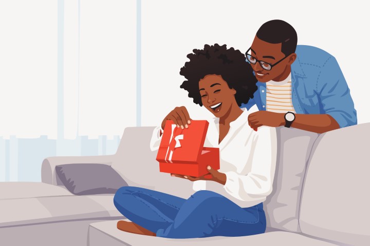 A woman sits on a sofa while her partner surprises her with a gift box.