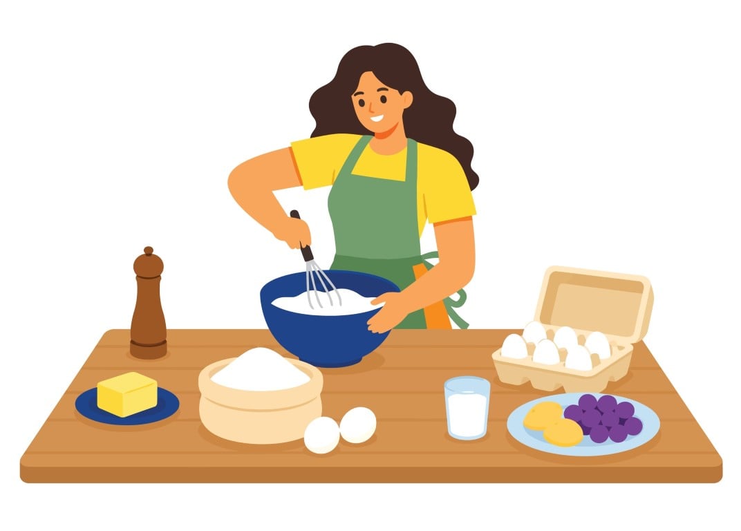 Smiling woman in an apron whisking batter at a kitchen table surrounded by baking ingredients.